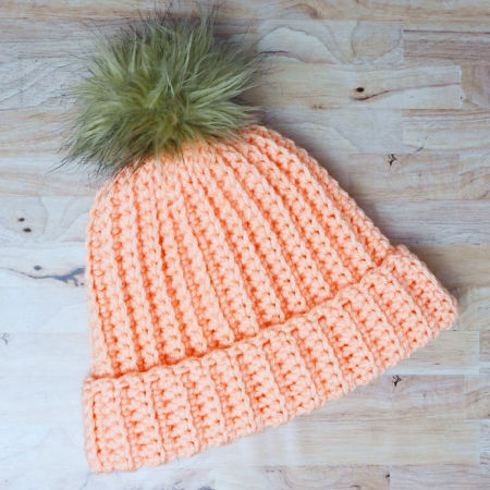 orange crochet beanie hat with a faux fur pom pom laying on a desk