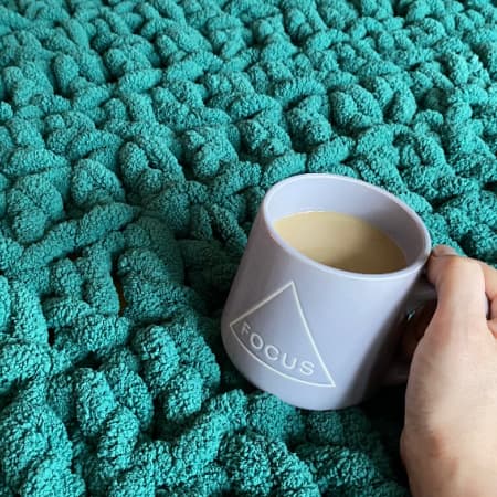 super bulky crochet throw blanket pattern layed out and I'm holding a purple coffee cup with coffee in it on top.