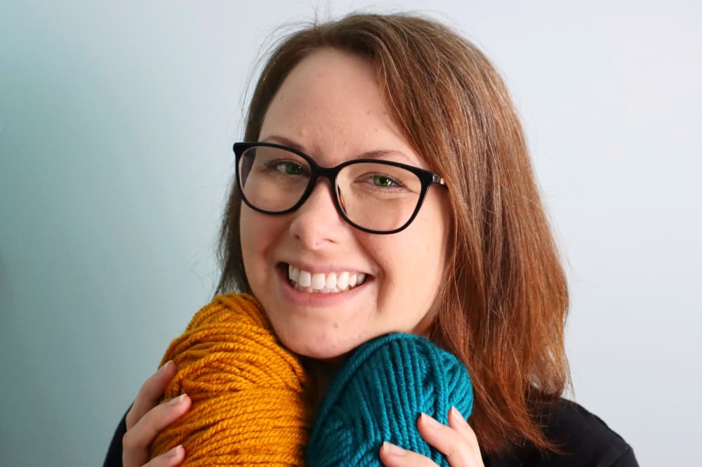 a picture of Crystal Martin from Marching North holding gold and teal yarn and smiling